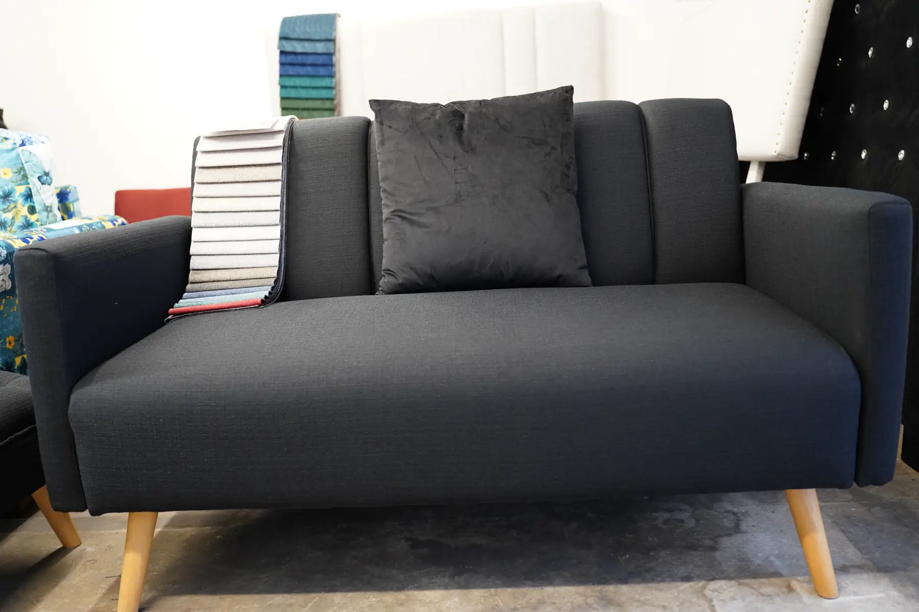 A charcoal sofa on pale timber legs with cushions and a fabric sample book on the arm.